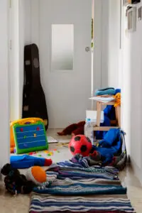 Professional organizer in Toronto A cluttered hallway featuring a door with a small window. Various children's toys, including a soccer ball, a colorful activity table, and clothing, are scattered on the floor along with a water bottle and a guitar case. offering professional organization services for homes and offices.