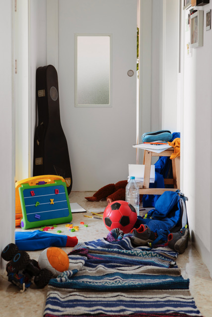 Professional organizer in Toronto A hallway cluttered with children's toys, including a yellow toy music box and a red soccer ball, alongside clothes and a water bottle, leading to a door with a frosted window. offering professional organization services for homes and offices.