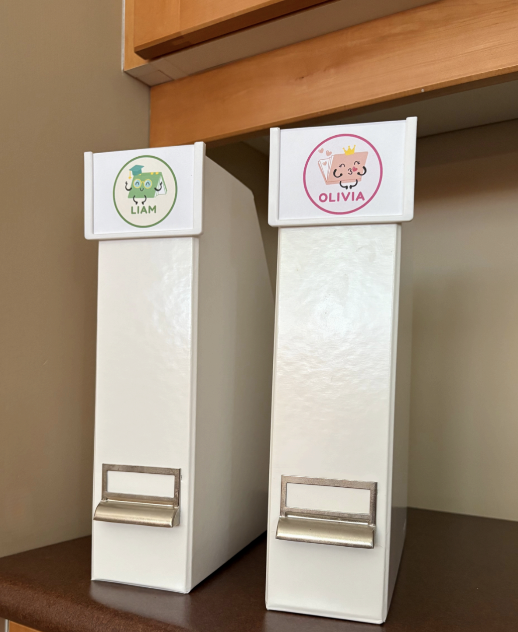 Professional organizer in Toronto Two white binders on a wooden shelf, labeled 'LIAM' with a green cartoon character sticker on the left, and 'OLIVIA' with a pink cartoon character sticker on the right. offering professional organization services for homes and offices.