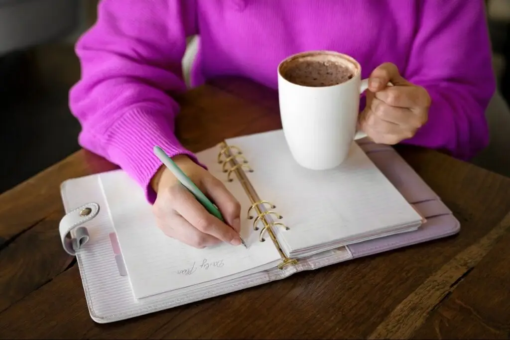 Professional organizer in Toronto A person in a bright pink sweater holding a white mug with a hot beverage, writing in a spiral-bound notebook on a wooden table. A green pen is in use, and the notebook is open to blank pages. offering professional organization services for homes and offices.