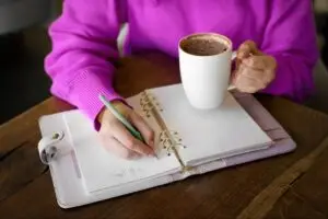 Professional organizer in Toronto A person in a bright pink sweater holding a white mug with a hot beverage, writing in a spiral-bound notebook on a wooden table. A green pen is in use, and the notebook is open to blank pages. offering professional organization services for homes and offices.