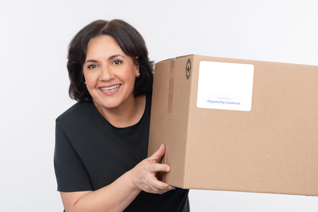 Professional organizer in Toronto A woman smiling while holding a large cardboard box with a label visible on it. She is wearing a black shirt against a plain white background. offering professional organization services for homes and offices.