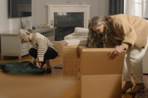 Professional organizer in Toronto Two women unpack belongings in a living room, with one kneeling over a green bag and the other lifting a cardboard box near a wicker chair and fireplace. offering professional organization services for homes and offices.